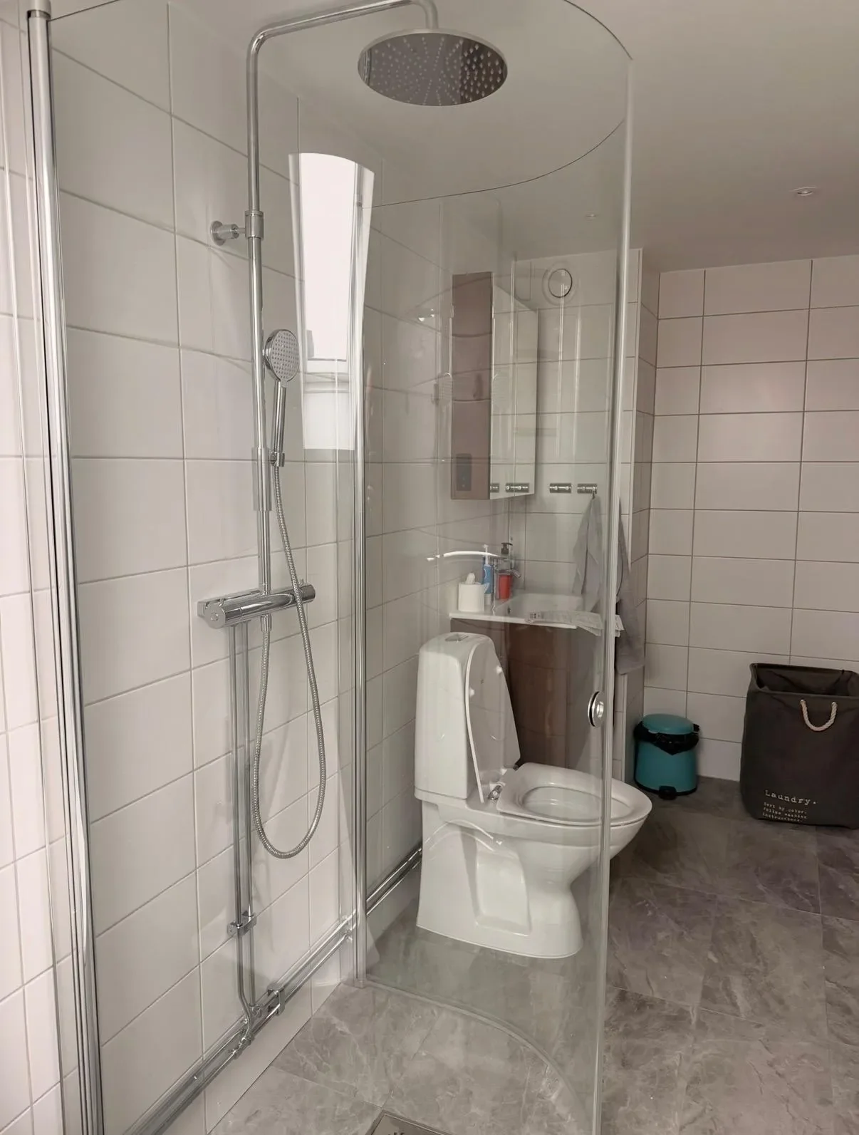 Vitt kaklat badrum med böjd glasdusch och ljusgrått klinkergolv