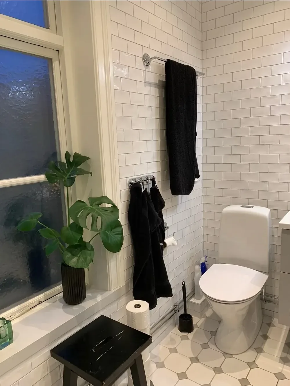 Badrum med vitt kakel, monstera vid fönstret och klinker i åttkantsmönster