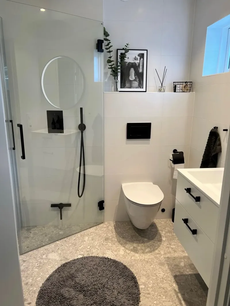 Vitt badrum med terrazzogolv, glasdörr till duschen och svarta blandare