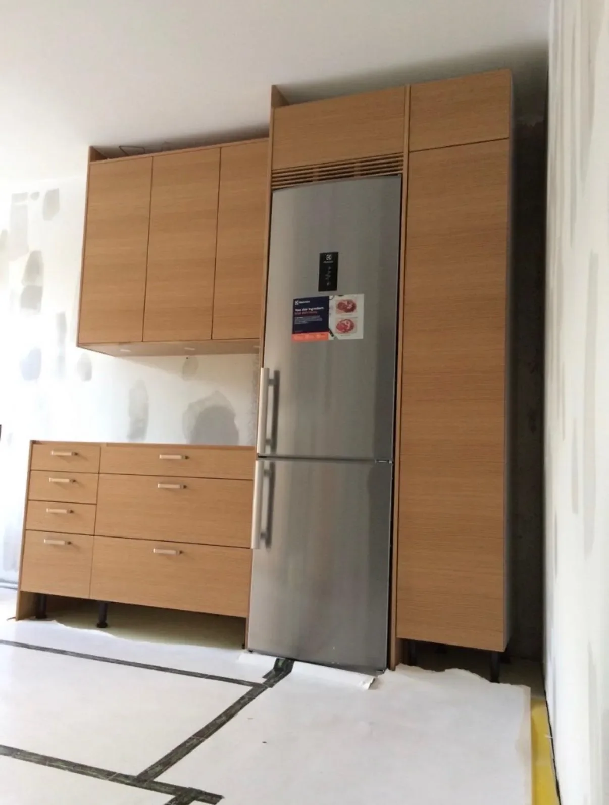 Kök under renovering med nya ekluckor, kylskåp på plats och skyddspapper på golvet