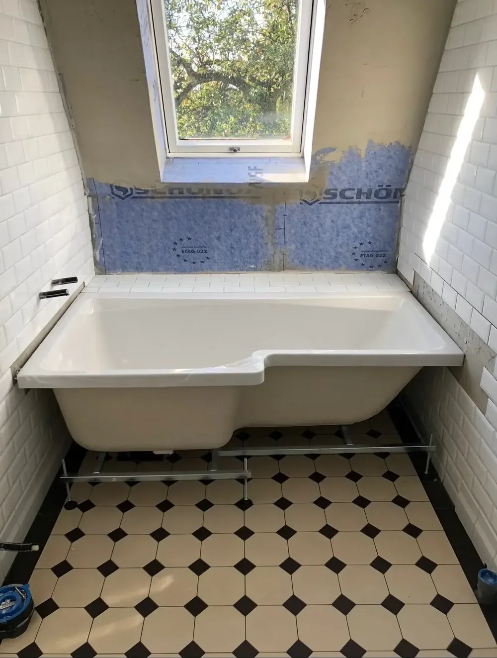Badrum under renovering med blått tätskikt på väggen bakom ett nymonterat badkar och åttkantigt klinkergolv